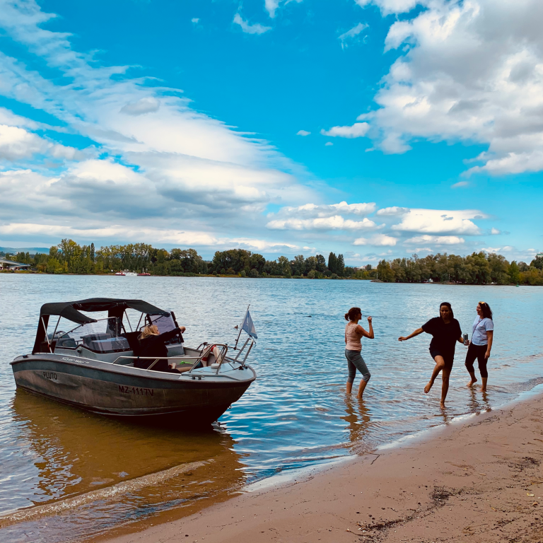 Drei Personen am Ufer eines Gewässers, ein Boot im Wasser und ein klarer Himmel.